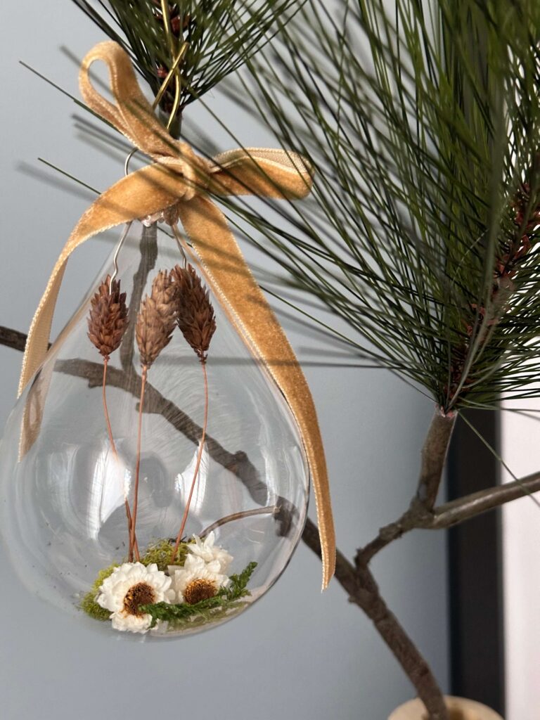 Glass ornament with dried flowers and a gold velvet ribbon hanging from a branch.