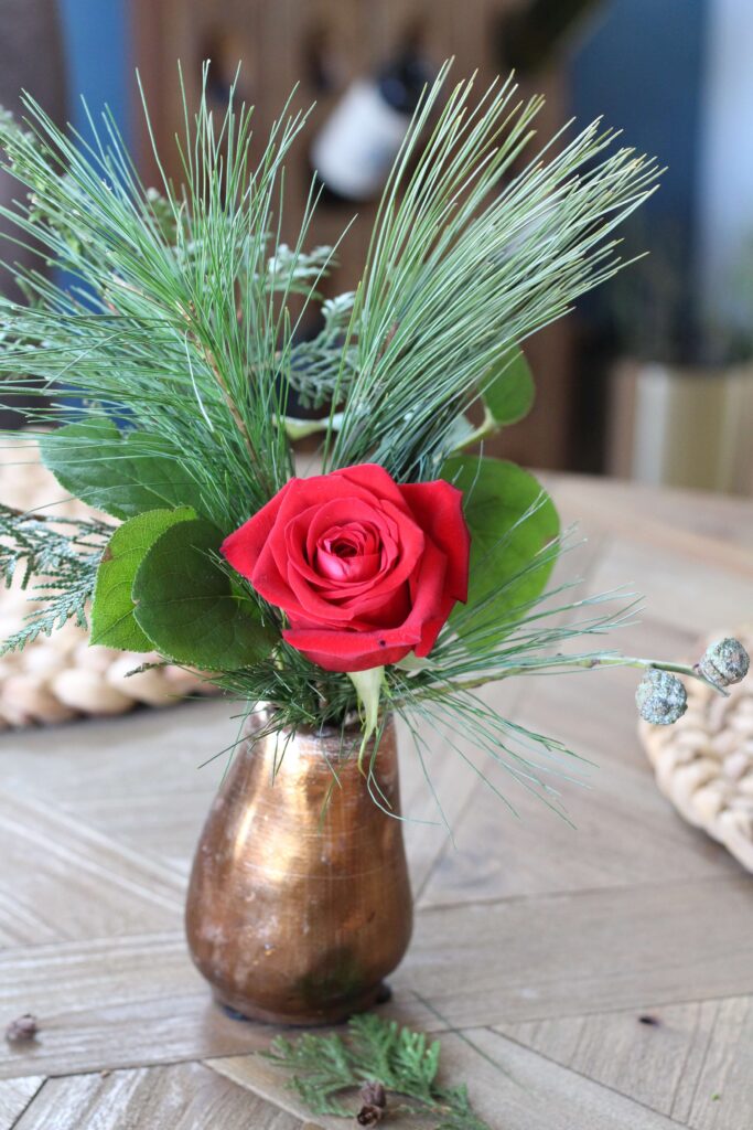 Small bronze bud vase with a red rose and sprigs of Christmas greenery.