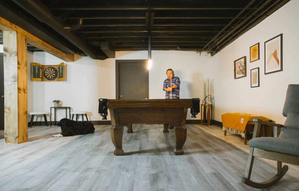 Basement remodel with black painted ceiling and peel and stick floor tiles and wood wrapped poles highlight a pool table.
