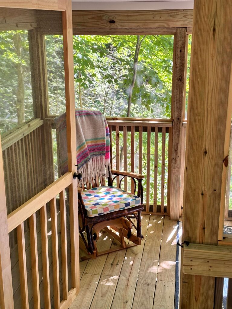 Wooden rocking chair with colorful cushions on a rustic cottage screened in porch.