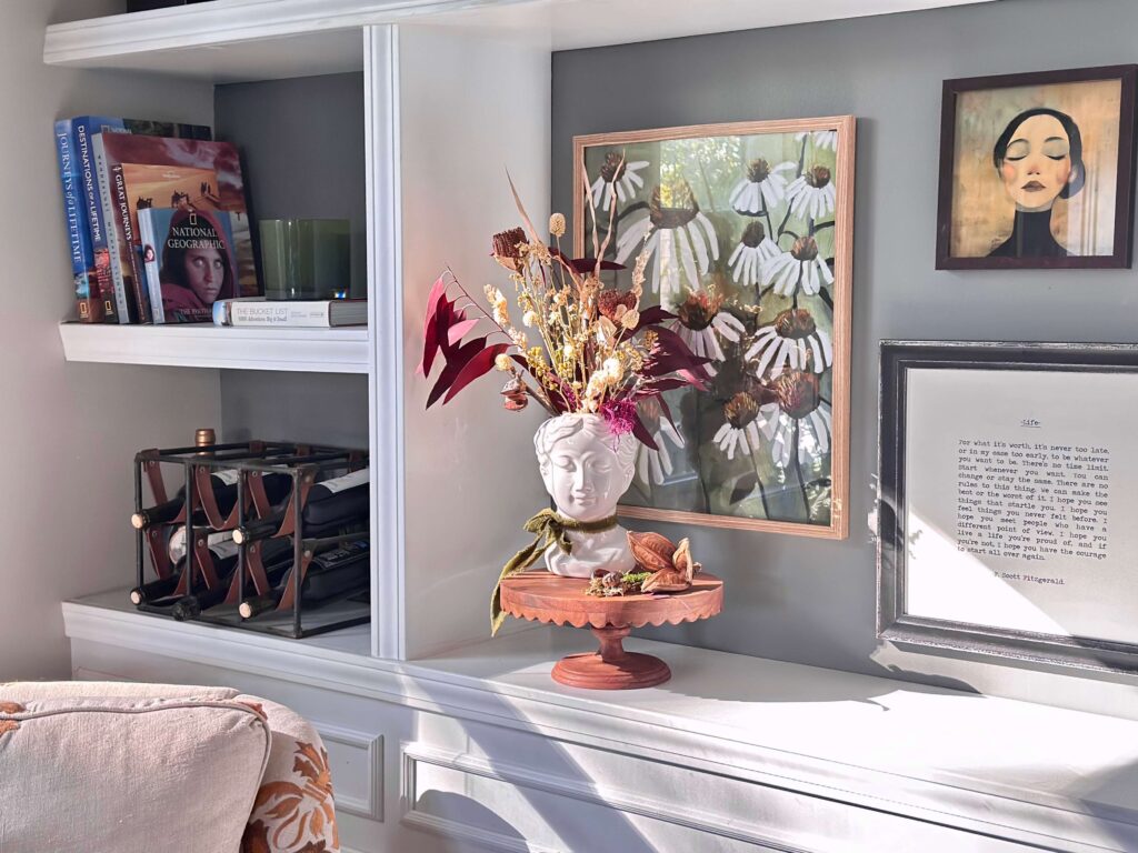 Bookcase with books, art and a Lady Head Vase filled with Dried flowers on a scalloped wooden cake plate.